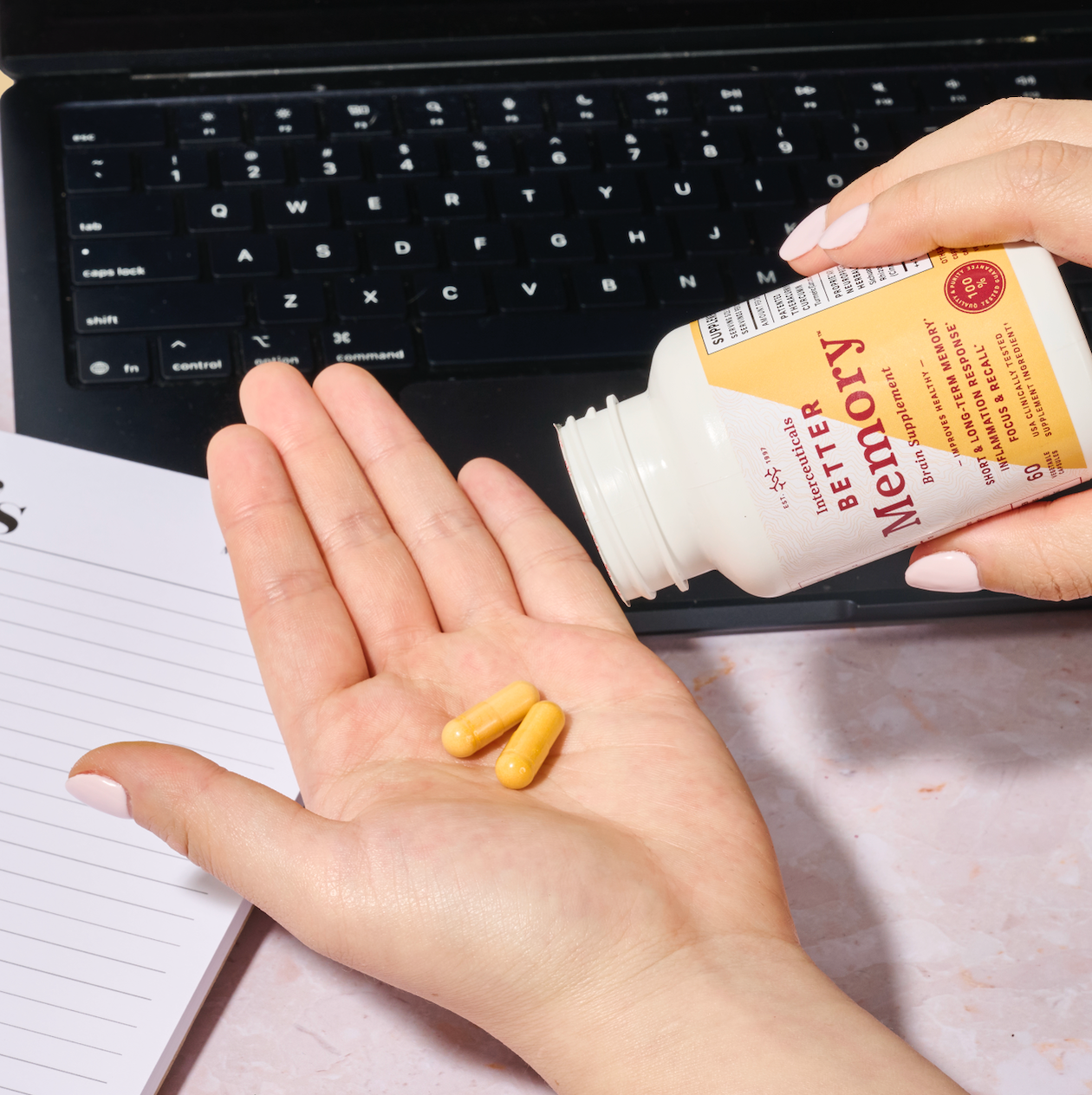 "Hand holding two yellow capsules and a bottle of Interceuticals Better Memory supplement, resting on a laptop keyboard and notepad, signifying a daily cognitive support routine.
